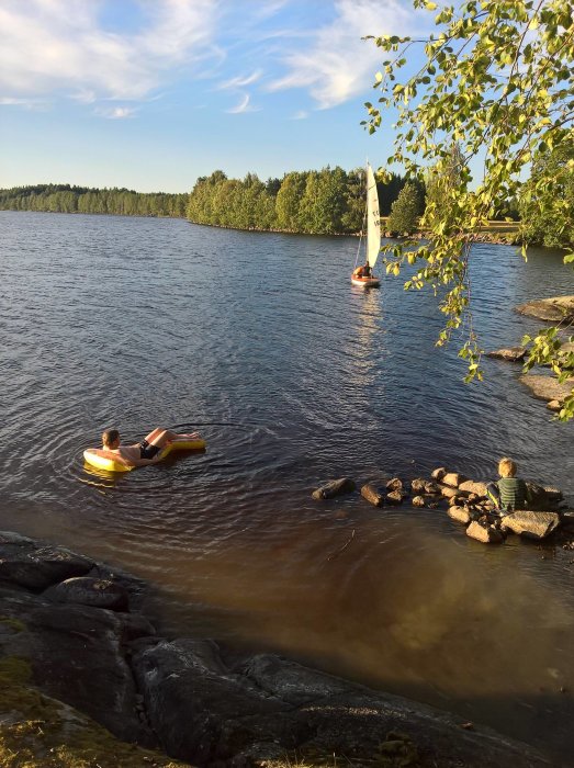 En badplats vid en sjö med en person som flyter på en uppblåsbar flotte och ett barn som sitter på stenar vid vattnet.