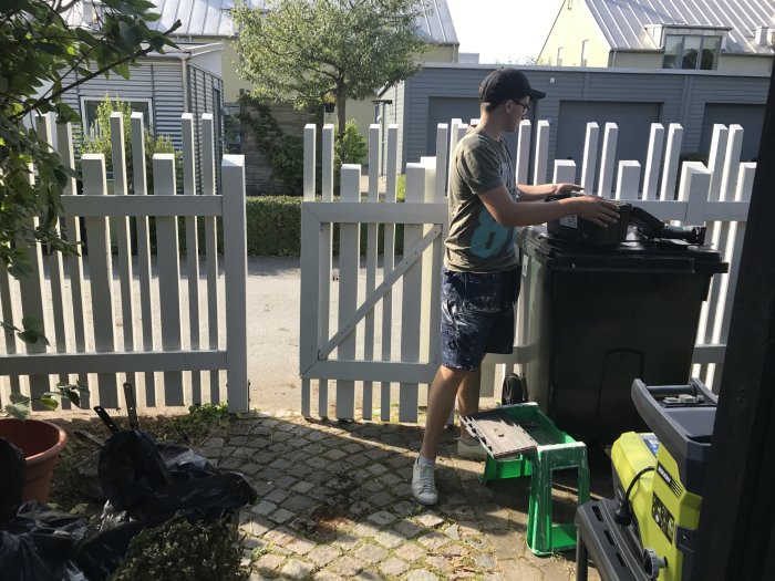Person i keps öppnar en nybyggd grind vid en uppfart med soptunnor i förgrunden.