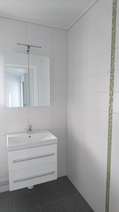 Renoverad toalett med vita kakelväggar, grå klinkergolv och modernt handfat med spegel.