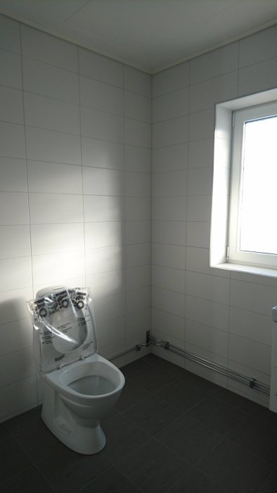 Nyrenoverad toalett med vita kakelväggar och mörkt klinkergolv, osättningsdetaljer synliga.