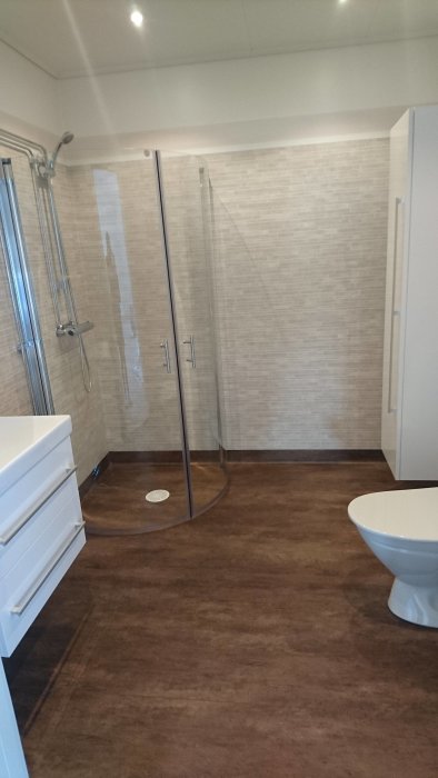 Renoverat badrum med beige kakelväggar och mörkt klinkergolv, duschhörna och toalett.