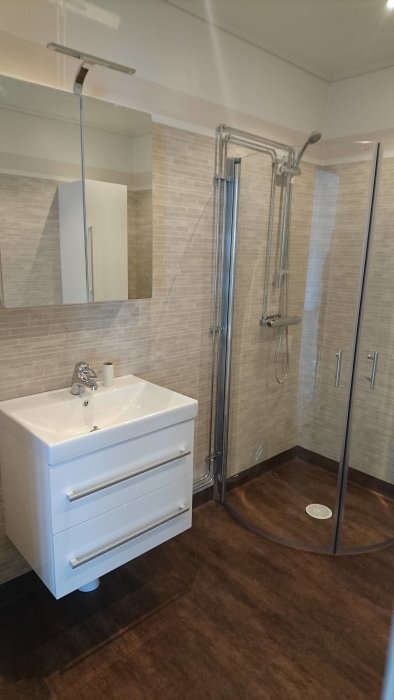 Nyrenoverat badrum med beige kakel, duschkabin och vitt handfat med skåp.