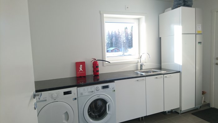 Nyinstallerad tvättstuga med vit tvättmaskin, skåp och diskbänk framför ett fönster med utsikt över snötäckt landskap.