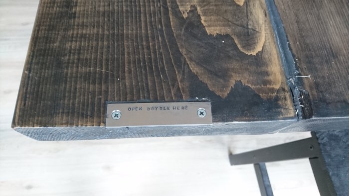 Hörnet av ett mörkt träbord med infälld metallplatta och texten "OPEN BOTTLE HERE".
