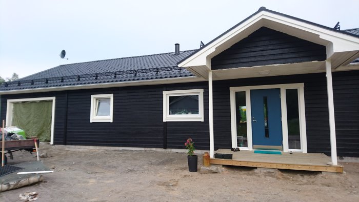 Nybyggt mörkblått hus med vita fönsterkarmar och en blå dörr, byggmaterial syns runtomkring.