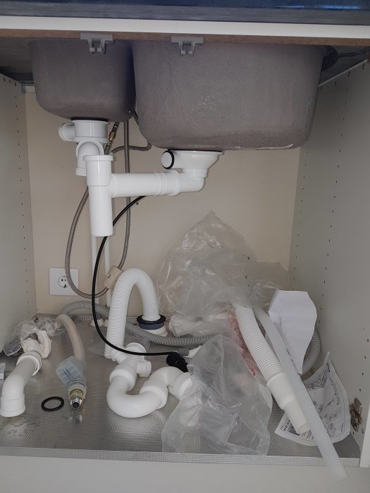 Oorganiserat köksskåp med avloppsrör, oanslutna slangar och byggmaterial under siligranit diskho.
