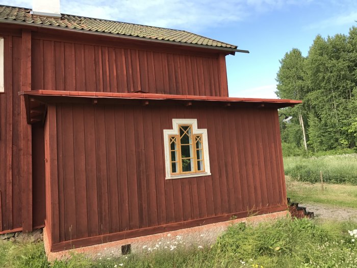 Rödfärgad träfasad på en byggnad med detaljerat takutspring och ett fönster med spröjs, omgivet av grönska.