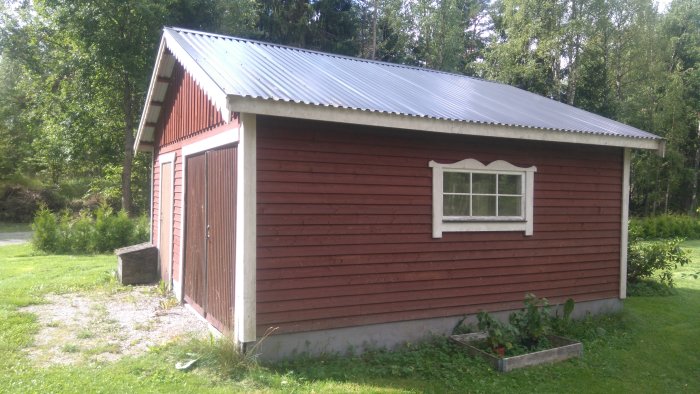 Rödmålad träkonstruktion av en garage/förrådsbyggnad med vitmålat fönster och plåttak omgiven av grönska.