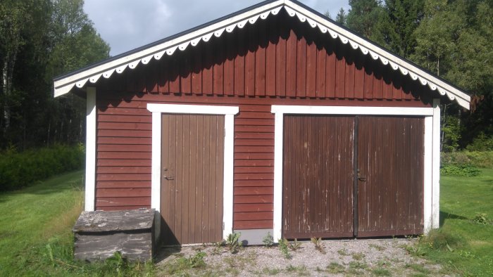 Ett rött garage med vit knut och två bruna dörrar under ett vitmålat tak med svarta takplåtar.