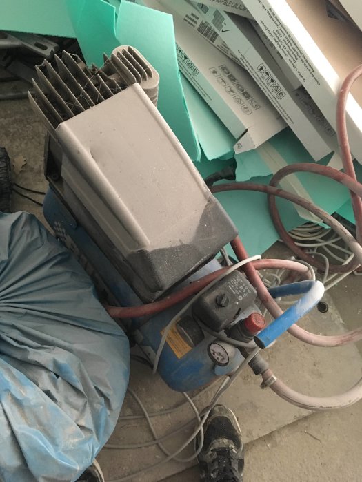 Bild av en dammig kompressor med slangar och kablar i en rörig verkstadsmiljö.