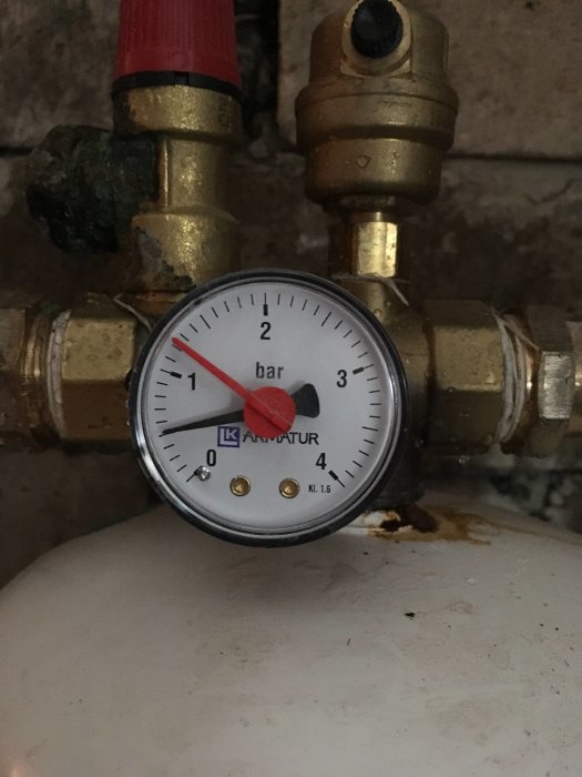 Tryckmätare med visare på 1 bar, monterad på rörledning ovanför en vit tank.