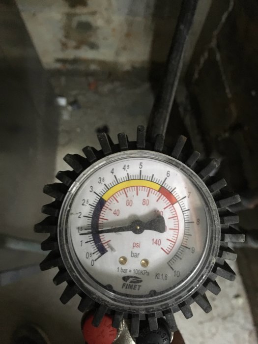 Manometer med visare som pekar på högt lufttryck, cirka 1,1 bar, i en verkstadsmiljö.