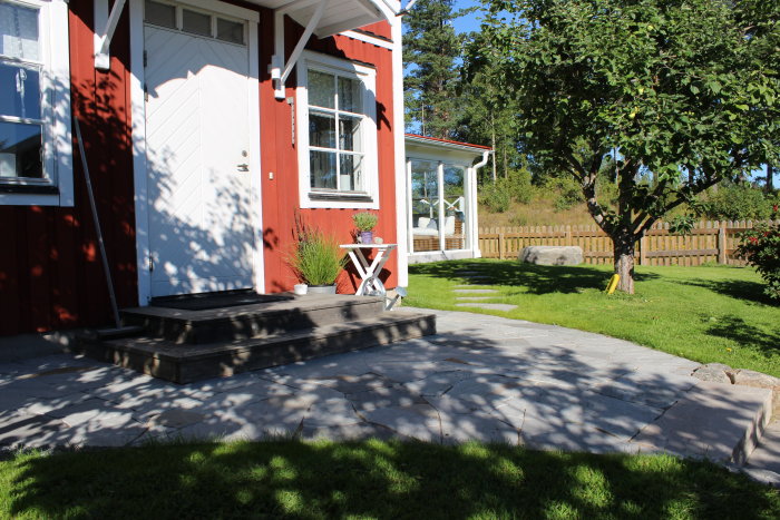 Tomt med nyväxt gräs, granitstolpar, sandstensplattor och stentrappor framför röd trähusentré.