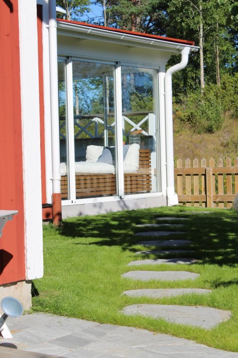 Stentrappa av sandsten leder upp till en vit inglasad veranda vid röd husgavel, omgiven av nylagt gräs och en trästaket.