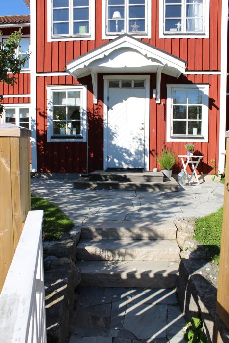 Renoverad tomt med gräs, granitstolpar, sandstensplattor och stentrappor framför rött hus med vita knutar.