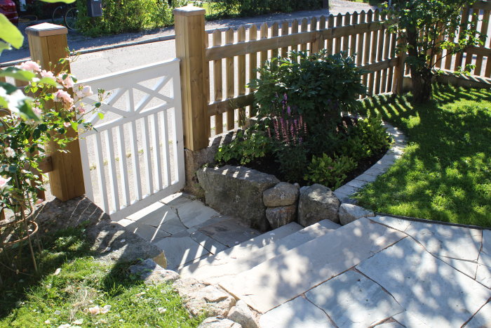 Trädgård med gräsmatta, blommande buskar och en stentrappa som leder till en vit grind och staket.