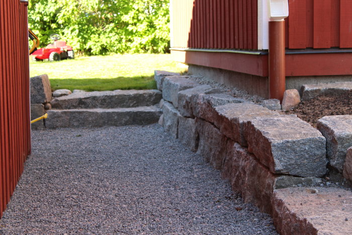 Nygräddat gräs med granitstolpar, stentrappa och tomtrabatter vid rött hus.