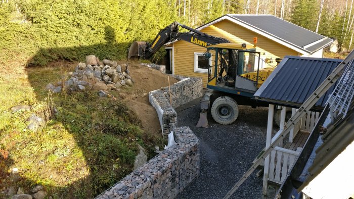 Grävmaskin vid husgrund under arbete med byggstenar och oanlagd trappa.