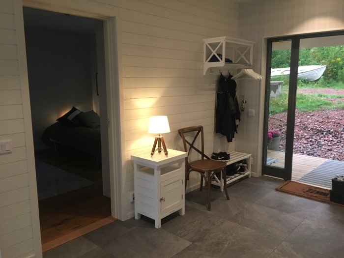 Invändigt hörn av ett hus med vit panel, en tänd lampa, en stol, och utsikt mot en öppen dörr och en trädgård.