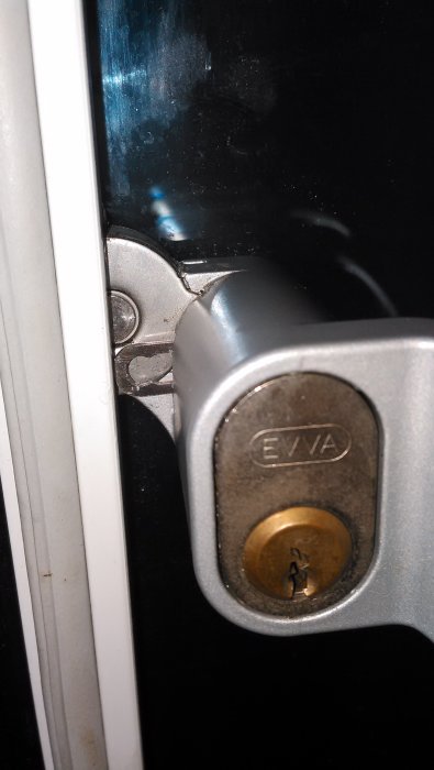 Oval låscylinder med kort medbringare monterad i dörrhandtag, visas med låsbricka och märkning EVVA.