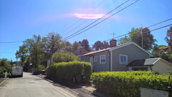 Solljus över en idyllisk gata med ett nyrenoverat ljusgrått hus och frodig häck.