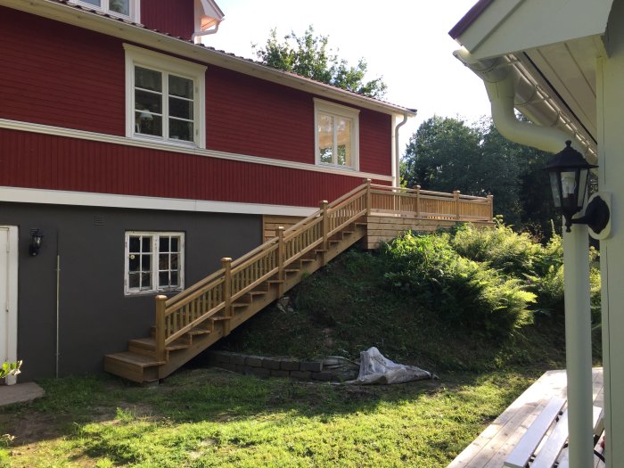 Nybyggd trätrappa och altan vid rödmålat hus med grön vegetation omkring.