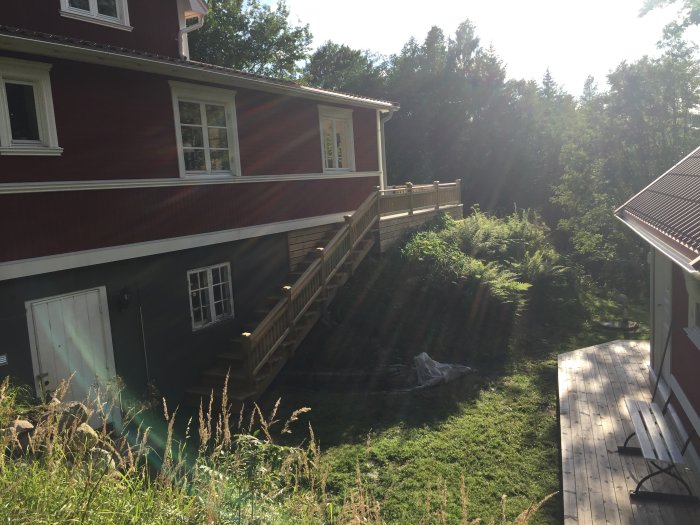 Solstrålar lyser genom träden på husets röda fasad med en nybyggd trappa som ansluter till en altan.