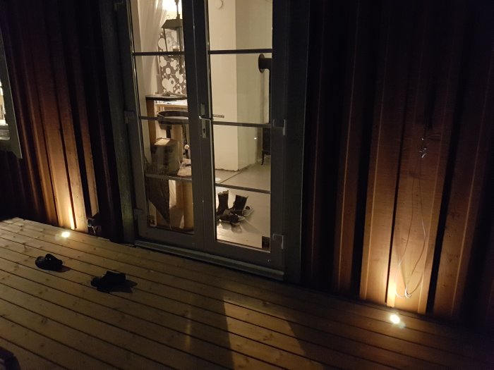 Altan med upplyst Cosa 45-inställningar på kvällen, par skor på trädäck, utsikt till interiör genom glasdörr.
