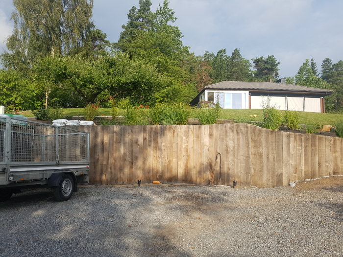 Sliten trästaket framför hus med trädgård, släp lastad med säckar parkerat till vänster.