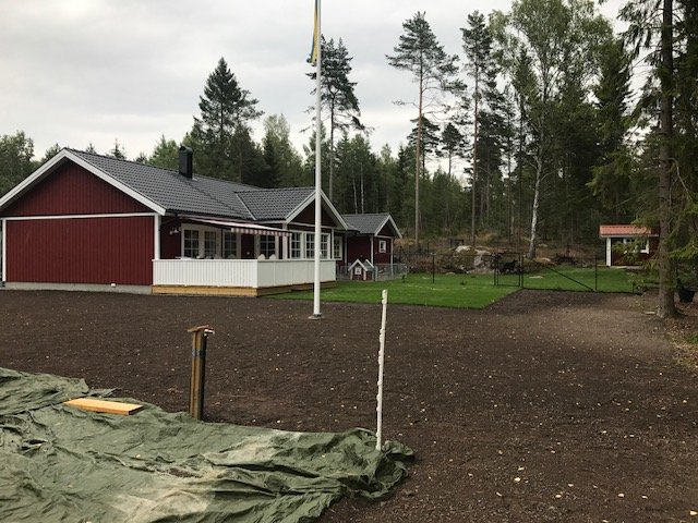 Rödmålat hus med flaggstång och staket, rullgräs, vedförvaring och pressenningtäckt trädgårdsland.