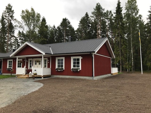 Rödmålat hus med vit knut, grusuppfart, flaggstång och skogsbakgrund, nyetablerat gräs på marken.