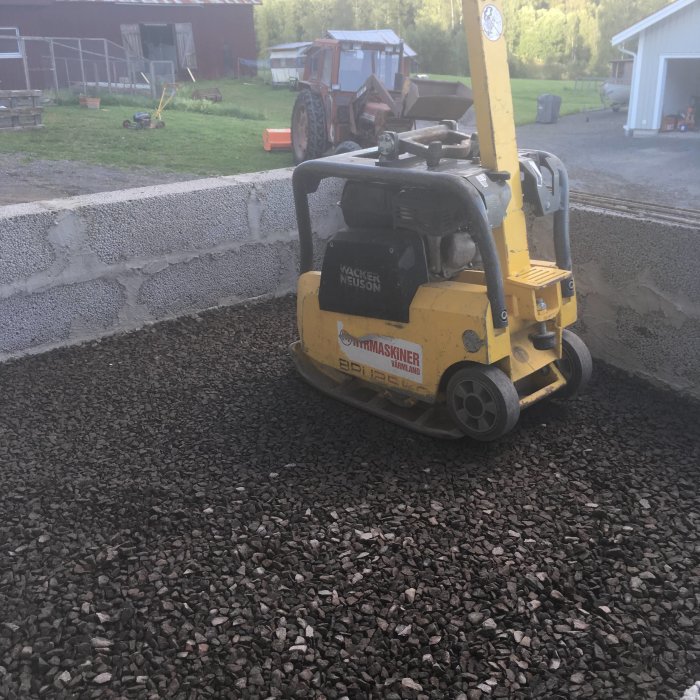 Gul markvibrator från Wacker Neuson vid en nylagd grusyta med byggmaterial och en traktor i bakgrunden.