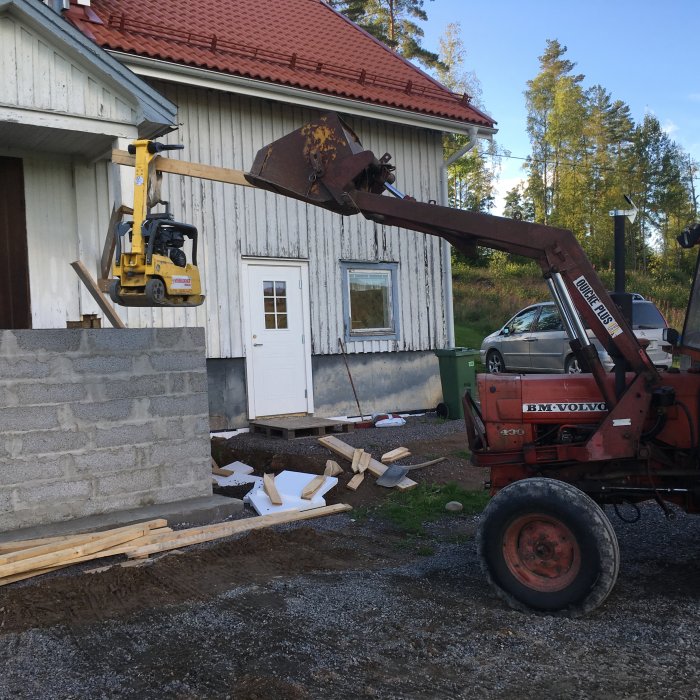 Renoveringsarbete med grävmaskin framför äldre hus, grusgrund och byggmaterial synliga.