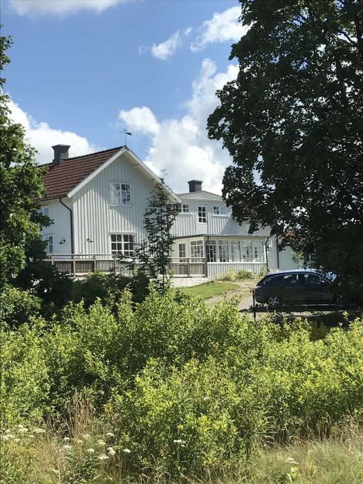 Vitt traditionellt hus med röda takpannor, omgivet av gröna buskar och träd under en blå himmel.