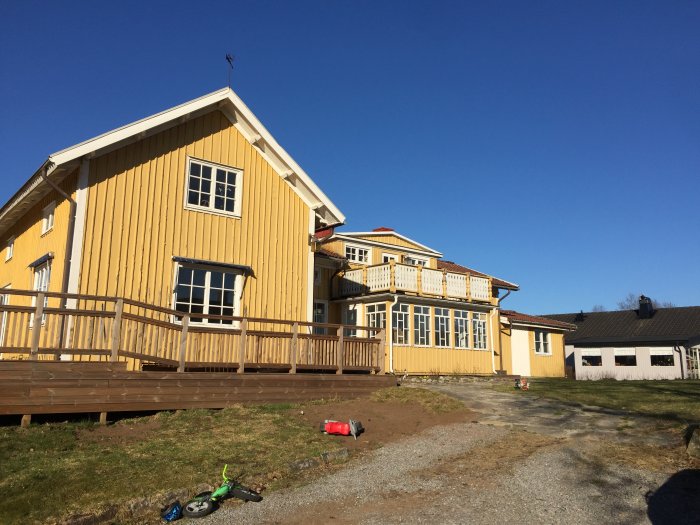 Gult trähus med veranda och barnleksaker på gården under klarblå himmel.