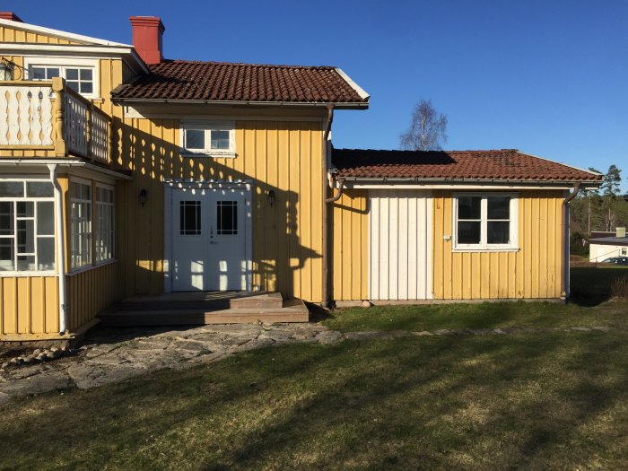 Gult trähus med rött tak och vit veranda i soligt väder.