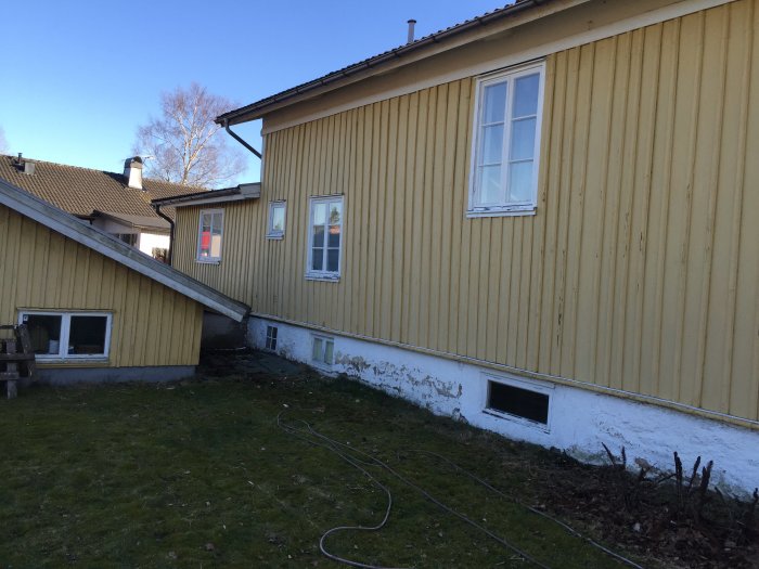 Gulmålat hus med vita fönster och slitet träverk, mot bakgrund av klarblå himmel.