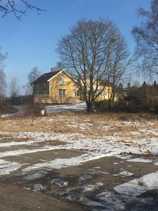 Ett tvåvåningshus målat i gult, med en glasveranda och en riven altan, omgivet av snötäckt mark och bar vegetation.