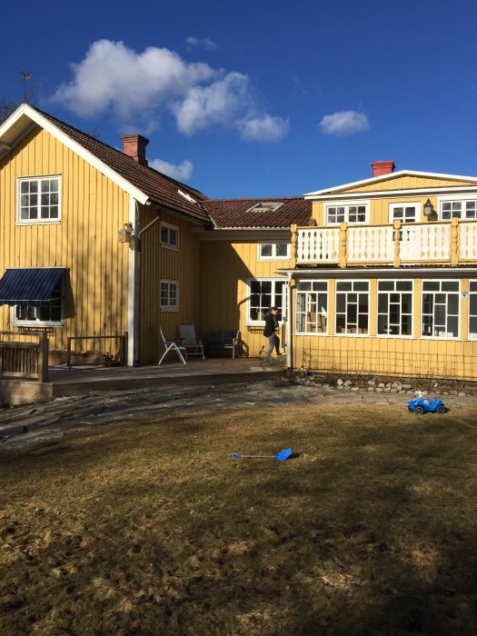 Gulmålat hus med glasveranda och saknat räcke, altan syns framför, grönt gräs och klarblå himmel.