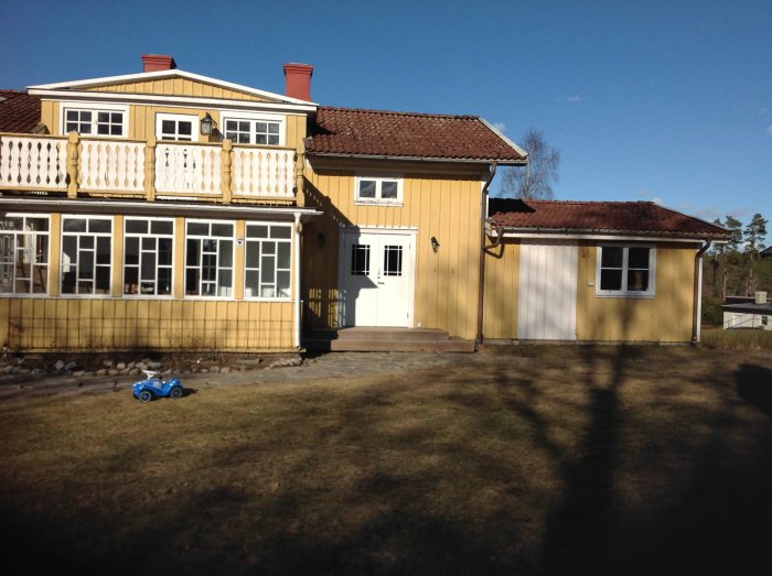 Gulmålat hus med glasveranda och saknat räcke, stora fönster och en leksaksbil på gräsmattan.