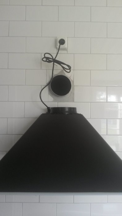 Svart köksfläkt monterad under kaklad vägg med synligt eluttag och rör som leder uppåt.