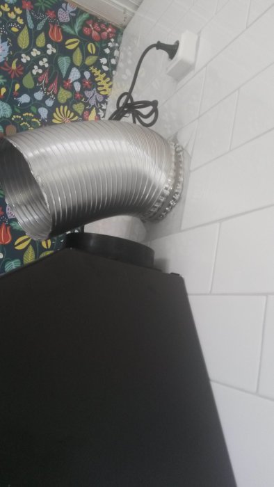Flexibelt rör i metall anslutet till en svart apparat, mot bakgrund av färgglad textil och vita kakelplattor.