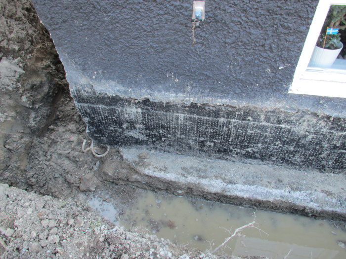 Skadad husgrund med synlig betongsula och vattensamling, före reparation.