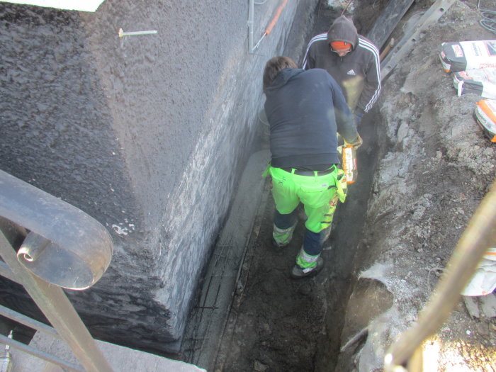 Två personer arbetar i en grävd grund vid husvägg, utför reparation av trasig sula med verktyg.