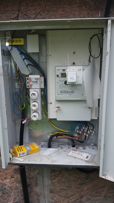 Öppet elkopplingsskåp med flera ledningar, automatsäkringar och en elmätare, med en tom förpackning vid skåpets botten.