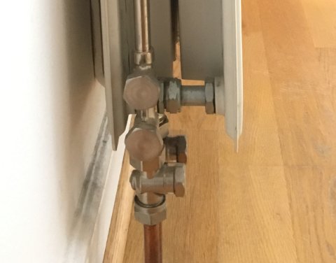 Radiatoranslutning med termostat och rör i en nisch vid trägolv och vit vägg.