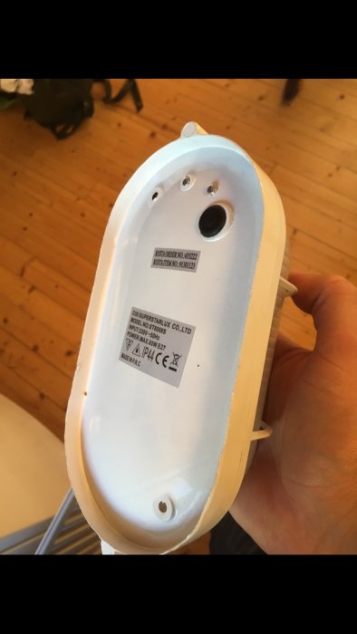 Baksidan av en vit plåtbyggnadslampa hållen i en hand, med etiketter och monteringshål.