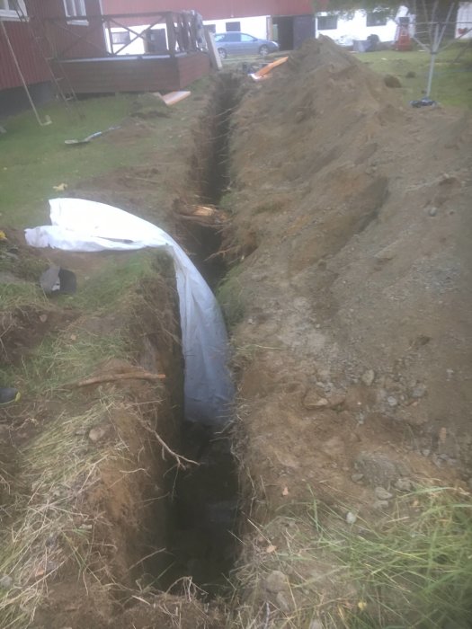 Nygrävd, lång grävning intill ett bostadshus med plastduk och uppkastad jord.