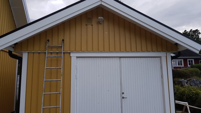 Gavelvy av ett gulmålat garage med vit garageport och en stege lutad mot väggen.