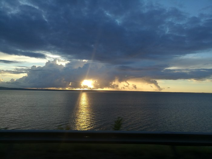 Solstrålar tränger genom moln ovanför en vattenspegling vid solnedgång.